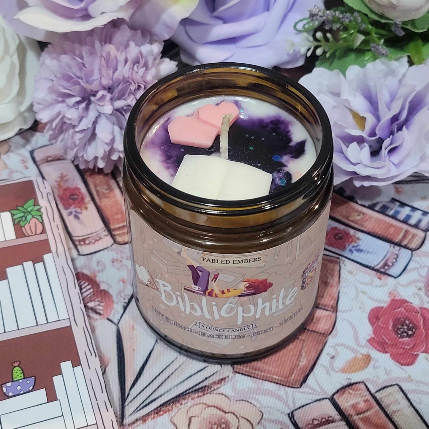 this photo showcases a cozy, bookish aesthetic featuring a beautifully crafted candle named "Bibliophile" from Fabled Embers.The candle is housed in a warm amber glass jar with a label that displays a charming illustration of books, a teacup, and a cozy reading scene. The wax inside has a mesmerizing galaxy-like swirl of deep purple and white, topped with adorable wax melts in the shape of a pink heart and a white book.
Surrounding the candle is a soft and dreamy setup—pastel purple and white flowers add a