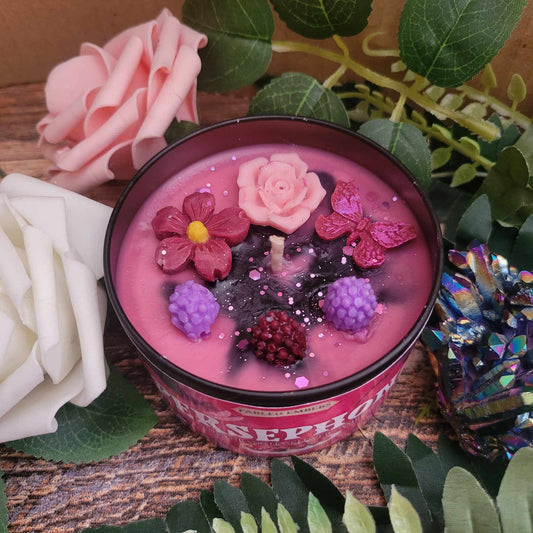 A beautifully crafted candle with a rich pink wax base sits surrounded by lush greenery and soft roses. The surface is adorned with intricate wax decorations, including a delicate pink rose, vibrant flowers, glittering pink butterflies, and realistic blackberries, with a hint of black swirling into the wax for a mystical touch. The label reads "Persephone", evoking themes of mythology, beauty, and mystery. The scene is set against a rustic wooden background, creating an enchanting and elegant aesthetic.