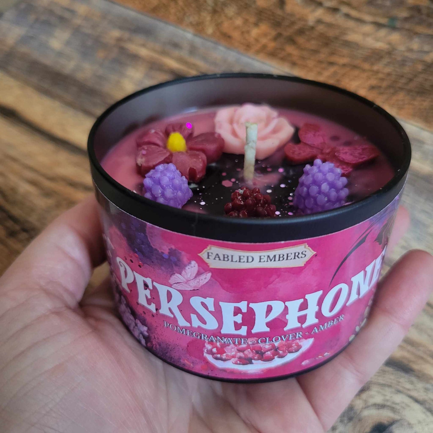 A beautifully crafted candle with a rich pink wax base sits surrounded by lush greenery and soft roses. The surface is adorned with intricate wax decorations, including a delicate pink rose, vibrant flowers, glittering pink butterflies, and realistic blackberries, with a hint of black swirling into the wax for a mystical touch. The label reads "Persephone", evoking themes of mythology, beauty, and mystery. The scene is set against a rustic wooden background.