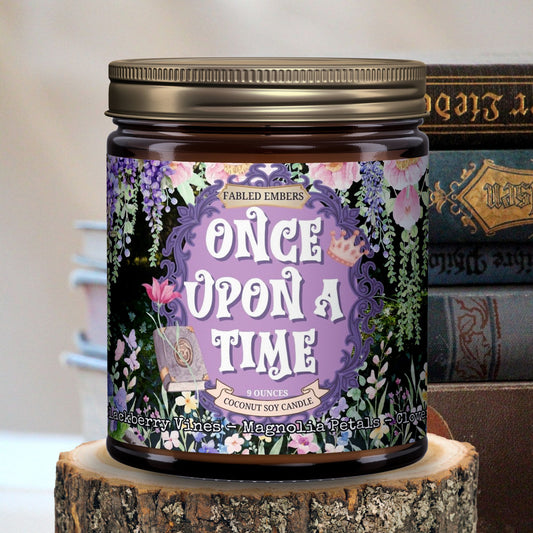 Amber jar candle with a purple label featuring an ornate frame, "Once Upon A Time" title, cascading pink and purple flowers, butterflies, and an open book. Scent notes include Blackberry Vines, Magnolia Petals, and Clover