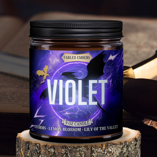 A jar candle with a black lid, labeled 'VIOLET' with a description of the scent profile including 'storms', 'earth', 'lily of the valley', and 'soft citrus', with a sword and book in the background