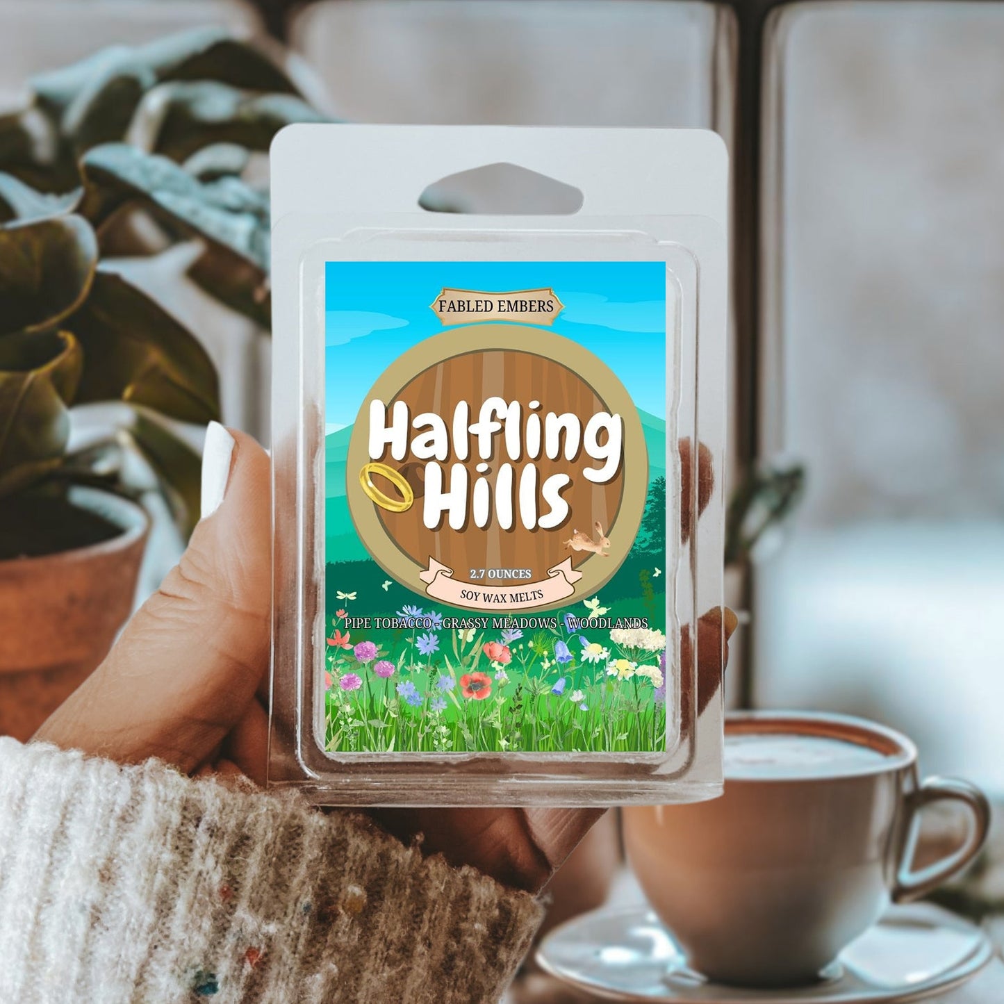 Hand holding a 'Halfling Hills' wax melt package with a blurred coffee cup and plant in the background