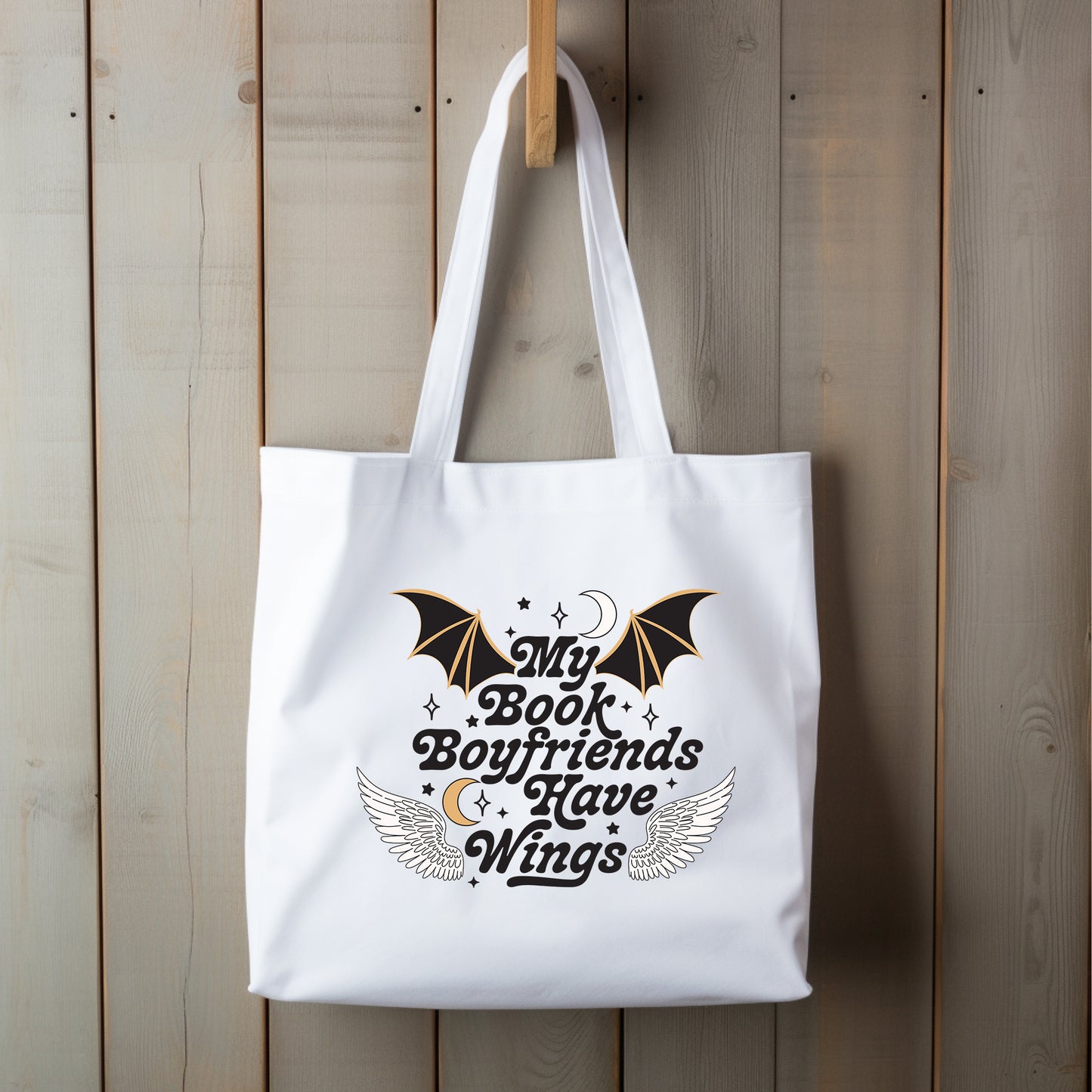 A white cotton canvas tote bag hanging against a wooden background, featuring text 'My Book Boyfriends Have Wings' along with wing graphics.