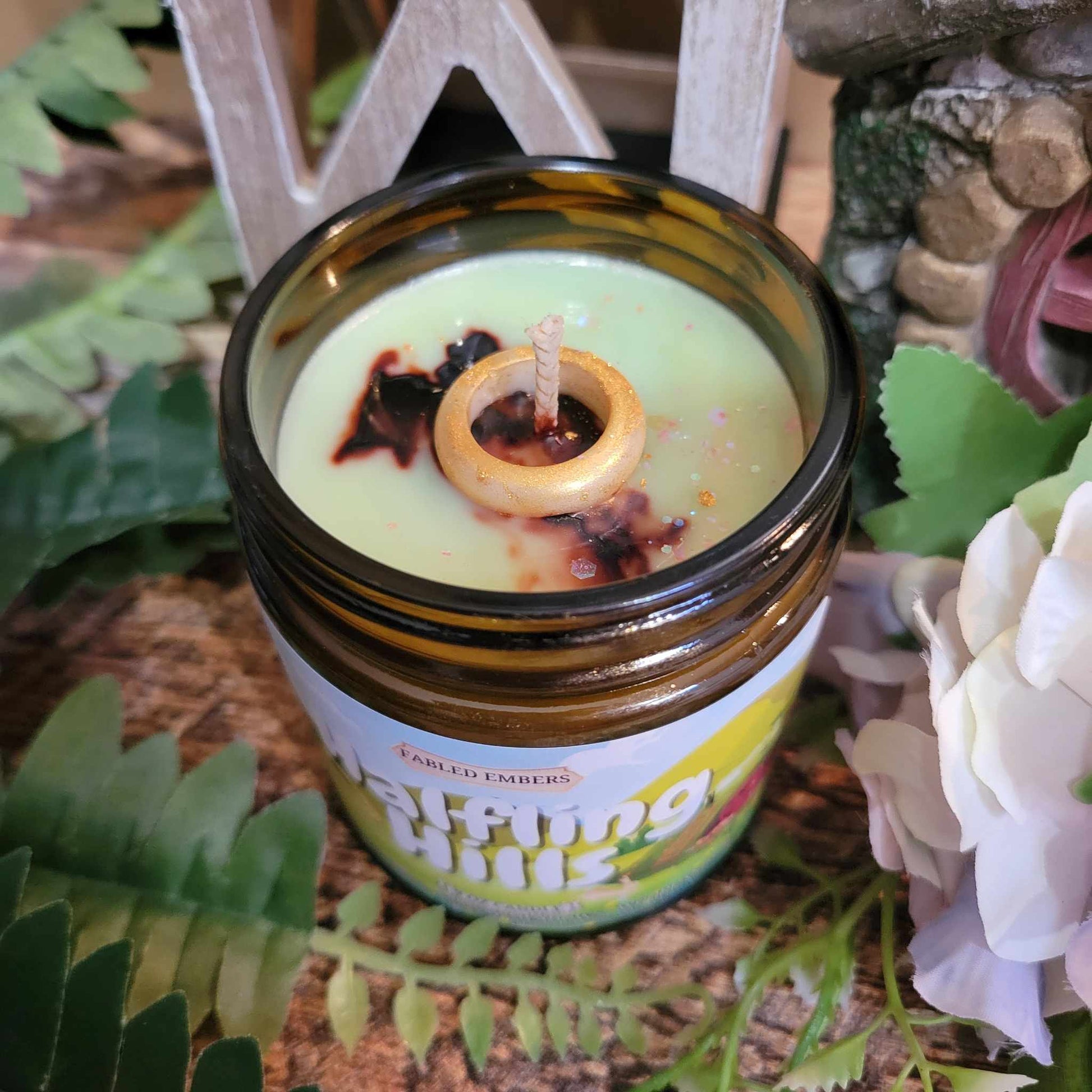 Candle in a glass jar with a wooden wick, surrounded by greenery and flowers.