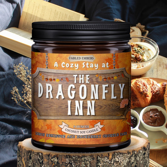 Candle labeled 'A Cozy Stay at The Dragonfly Inn' on a wooden block with a person reading a book in the background.