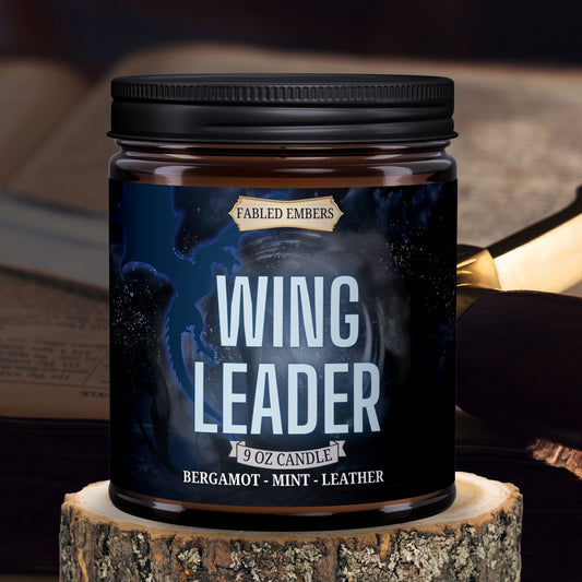 Wing Leader candle by Fabled Embers featuring  a blue dragon on a label with shadow moitiffs, amber glass jar sitting on a wood riser, with a sword and book in the background